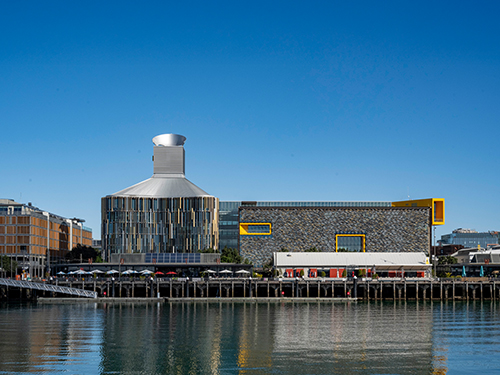 Waterfront view of a modern commercial building complex with distinctive architecture reflected in calm harbour waters under a clear blue sky. 