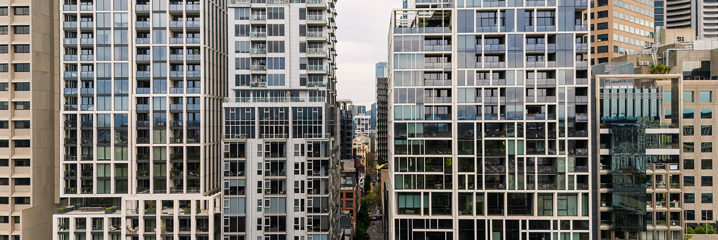 Modern high-rise residential buildings with glass facades and balconies closely arranged along a narrow city street in a dense urban environment.