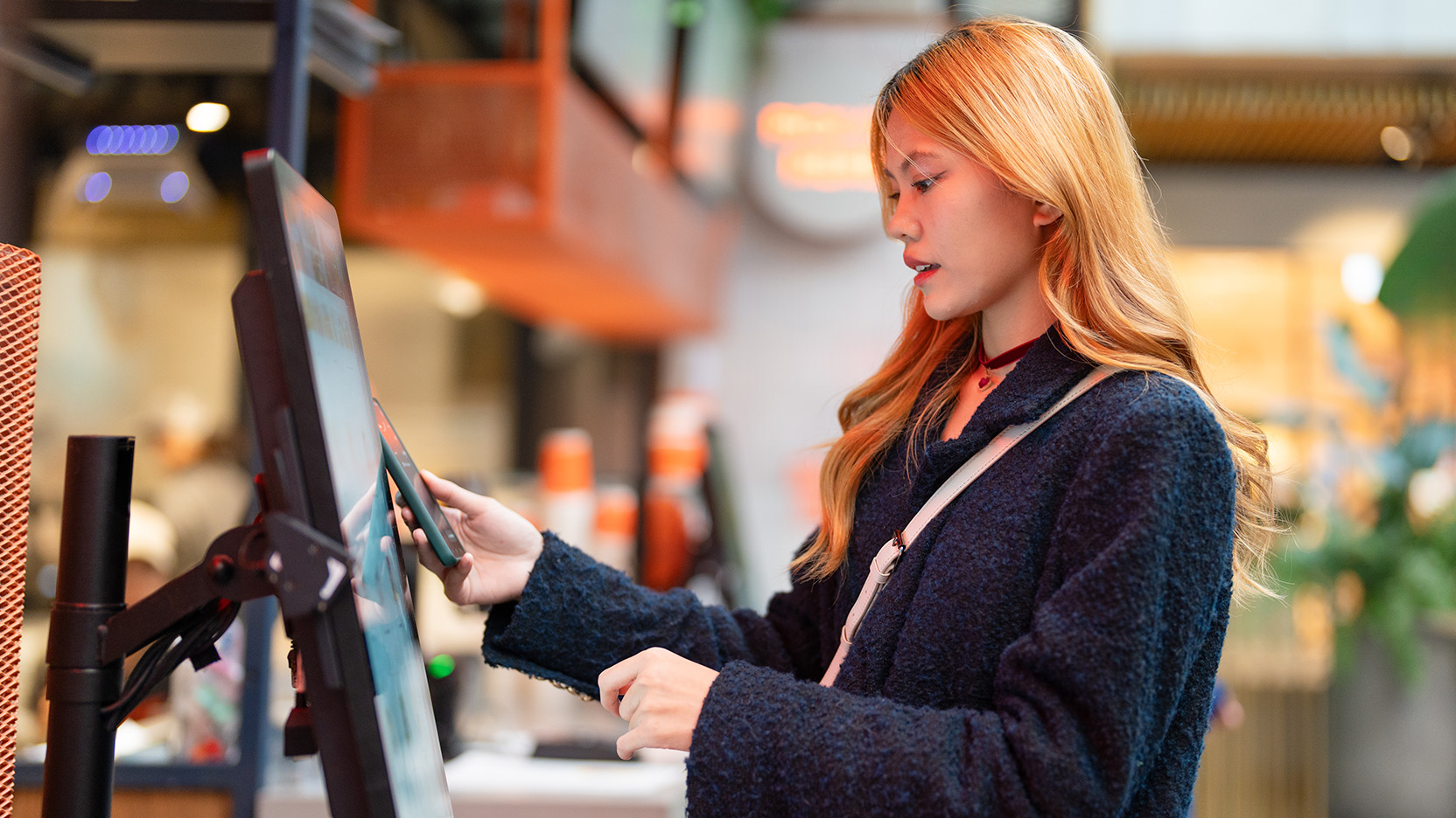 Person using a touchscreen self-service kiosk and smartphone in a modern quick-service environment. 