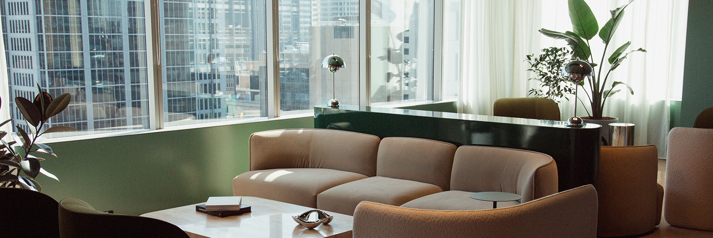 Modern office lounge with curved seating and a coffee table, set beside large windows overlooking a dense city skyline, with indoor plants and soft natural light.