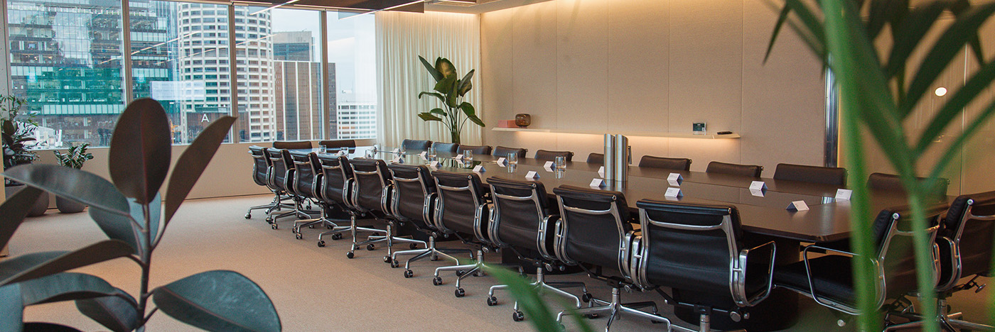 Modern conference room with a long boardroom table surrounded by black office chairs, set beside large windows overlooking a city skyline, with indoor plants and soft ambient lighting.