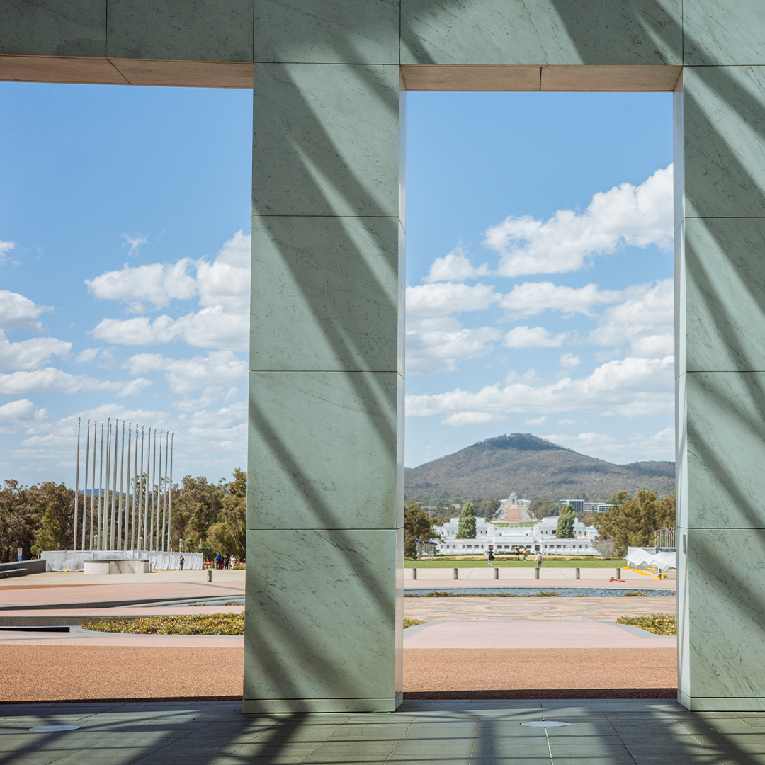 Modern civic plaza framed by monumental stone columns, with views towards a landmark government precinct and distant hills under a clear sky.