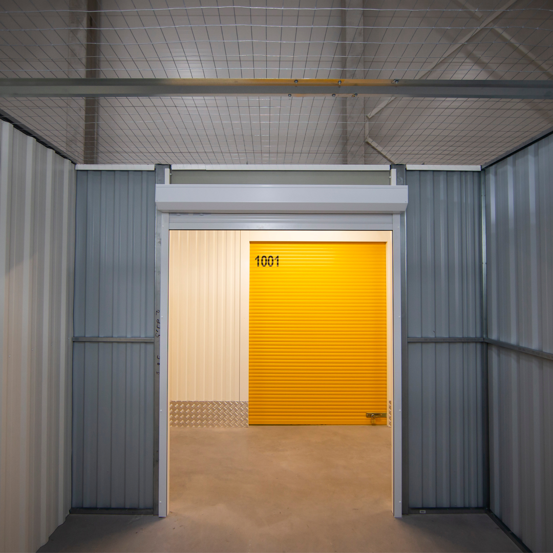 A bright yellow roller door marked "1001" is seen within a self-storage unit, with corrugated metal walls and a wire mesh ceiling.