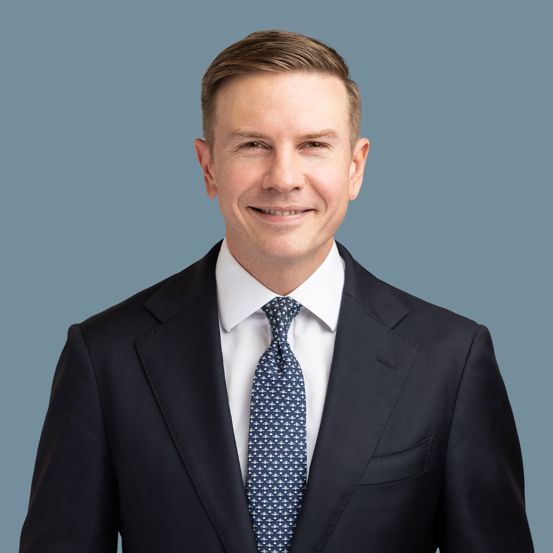 Professional headshot of a person in a dark suit and patterned tie against a plain blue background.