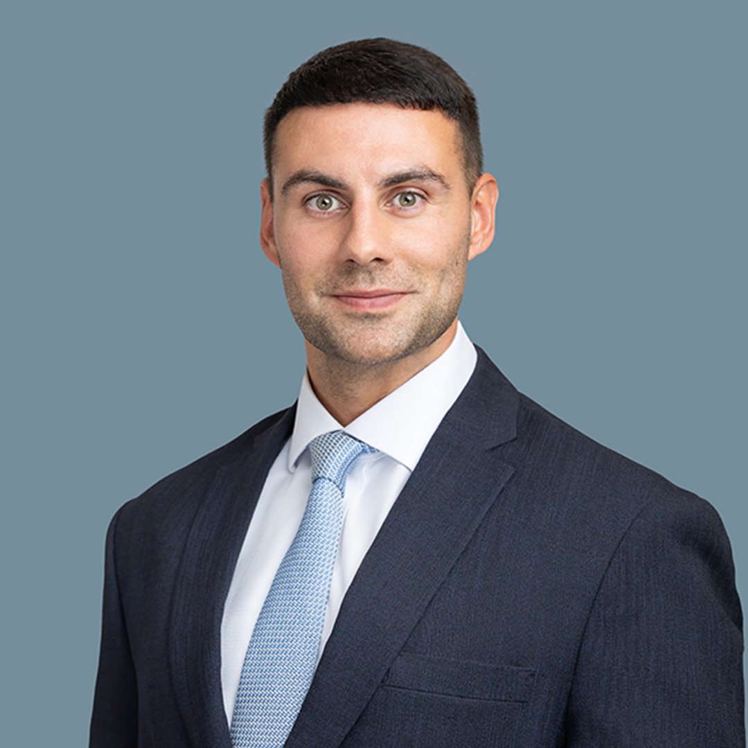 Professional headshot of Sam Saghir Pashley in a dark business suit and light blue tie against a plain blue-grey background. 