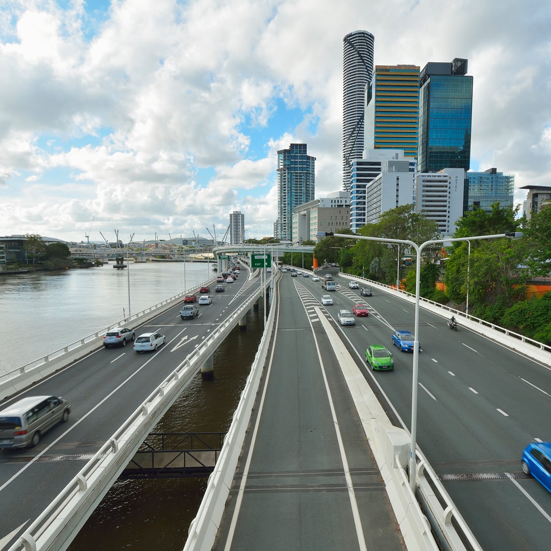 An aerial view shows a multi-lane highway bustling with cars, splitting around a river in a modern city with numerous high-rise buildings under a cloudy sky.