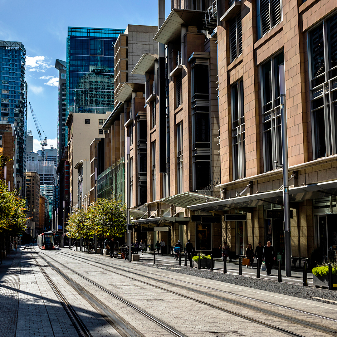 A light rail tram glides along tracks through a sunny Sydney city street lined with modern high-rise office buildings and pedestrians.