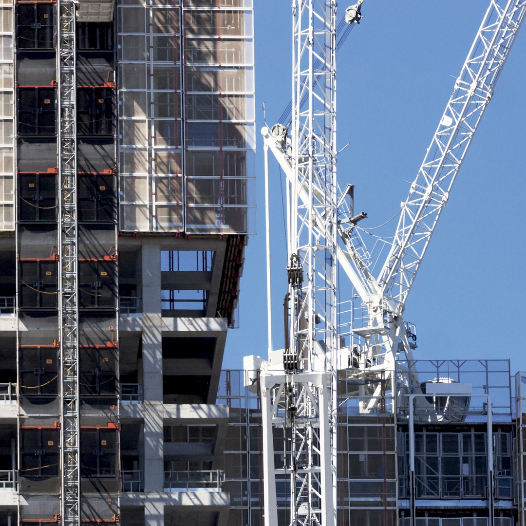 A tall, partially constructed building with scaffolding and a large white crane stands against a clear blue sky.