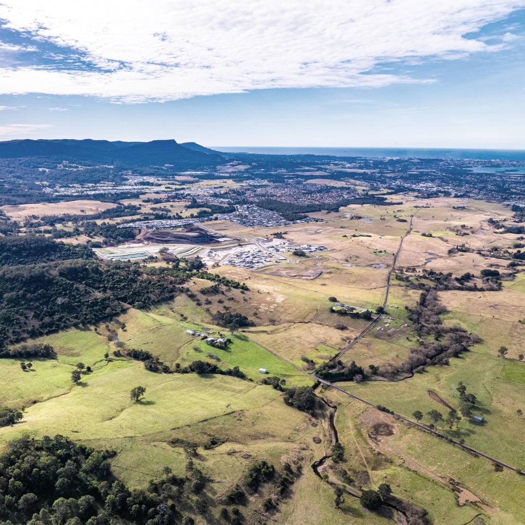 An aerial view looking over rolling green paddocks toward a suburban housing development and the distant coastline under a bright blue sky.