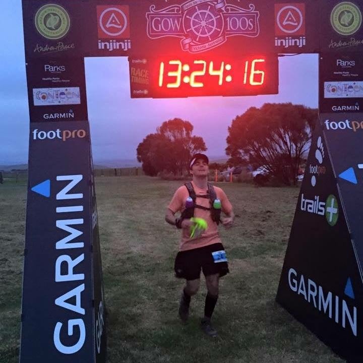 James King crosses the finish line of an ultramarathon beneath an illuminated race clock, wearing trail running gear during a long distance endurance event.