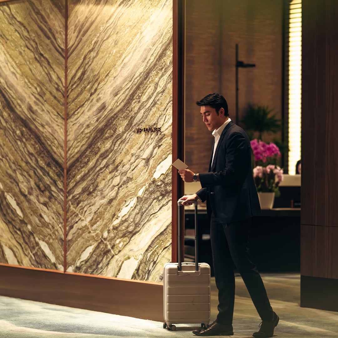 A hotel guest with rolling luggage walks through an upscale lobby featuring a dramatic stone feature wall, warm lighting and premium guest amenities. 