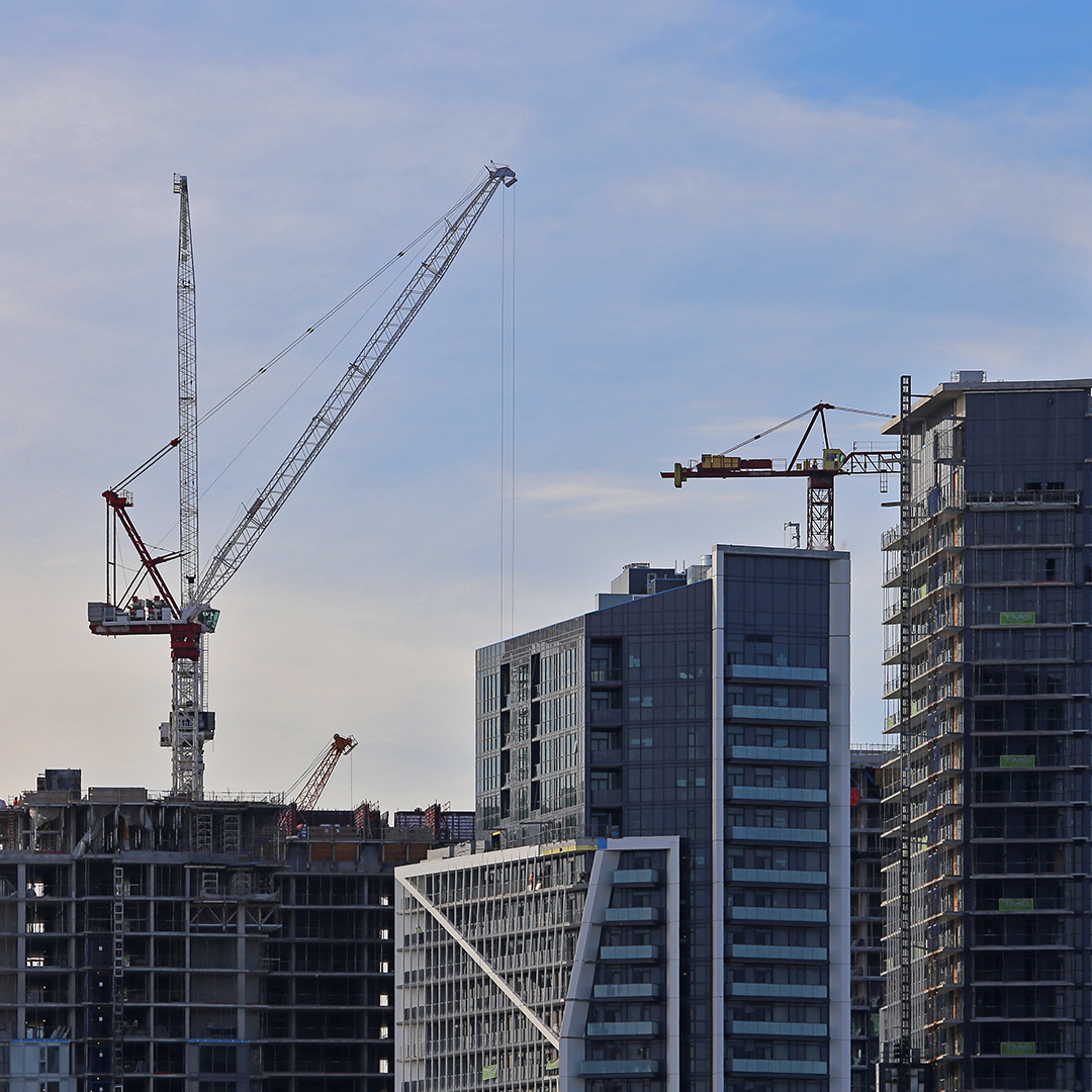 A busy construction site under a partly cloudy sky, with several cranes towering over a mix of finished and unfinished modern high-rise buildings.