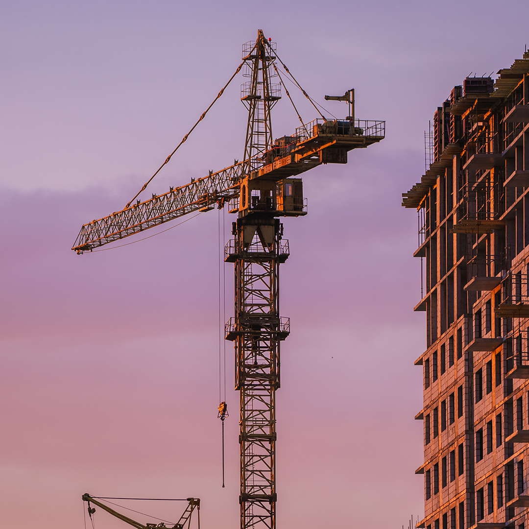 A tall construction crane stands next to a multi-storey building under construction, all bathed in the warm glow of a sunset sky.