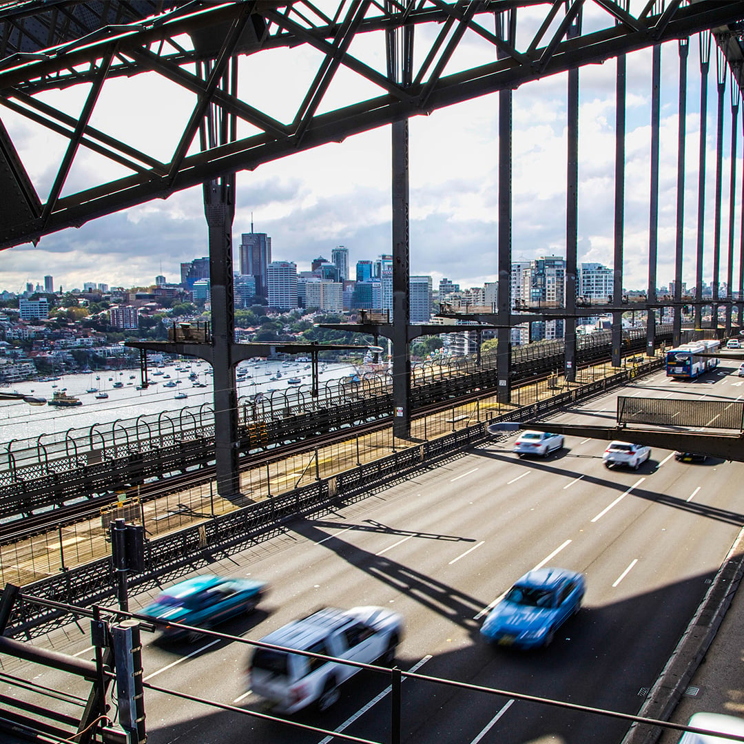Sydney's cityscape is seen through the steel framework of the Harbour Bridge, with cars speeding along the roadway below.