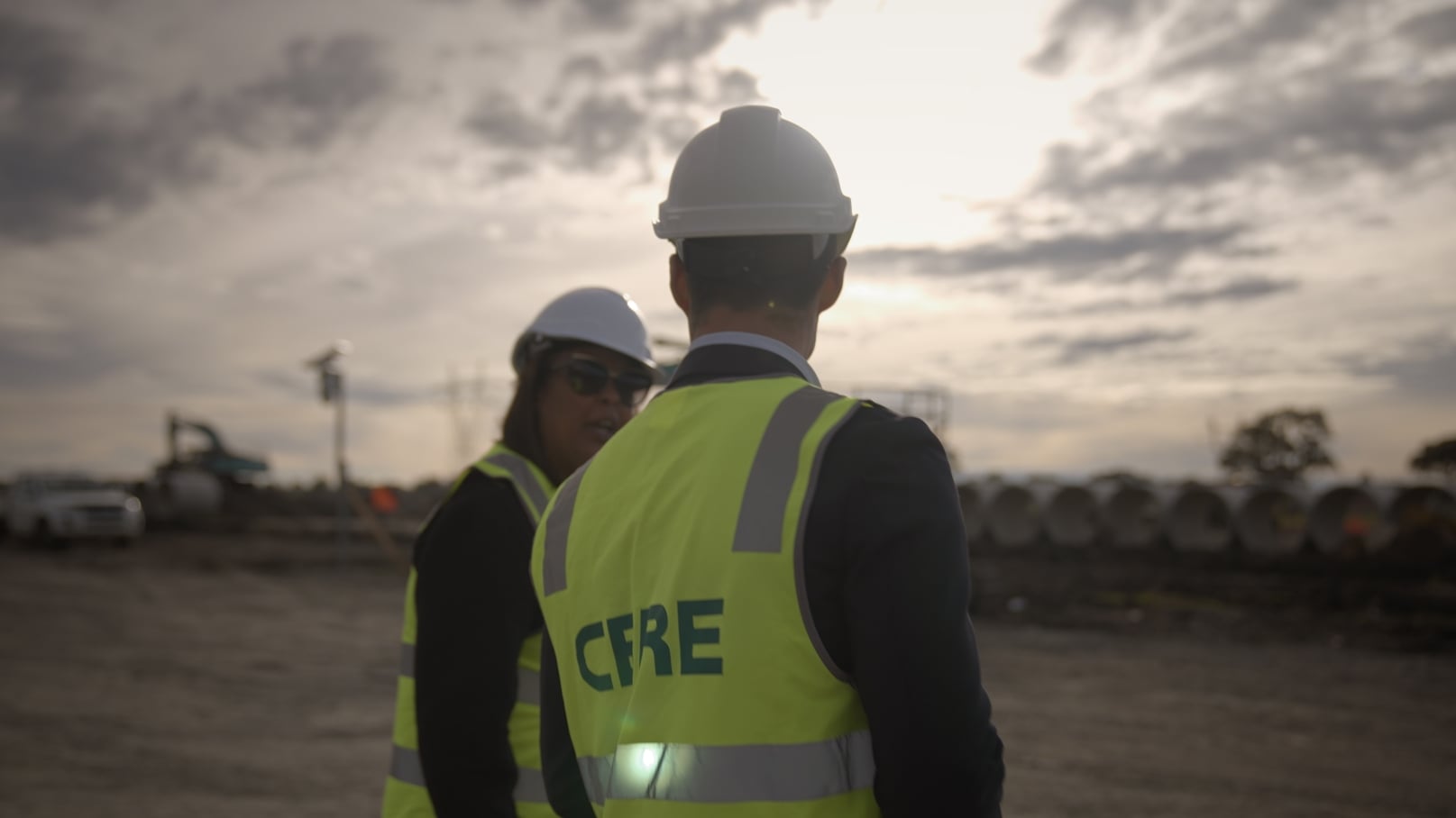 Two individuals wearing hard hats and high-visibility vests stand on a dusty, outdoor site with a cloudy sky and bright sunlight in the background.