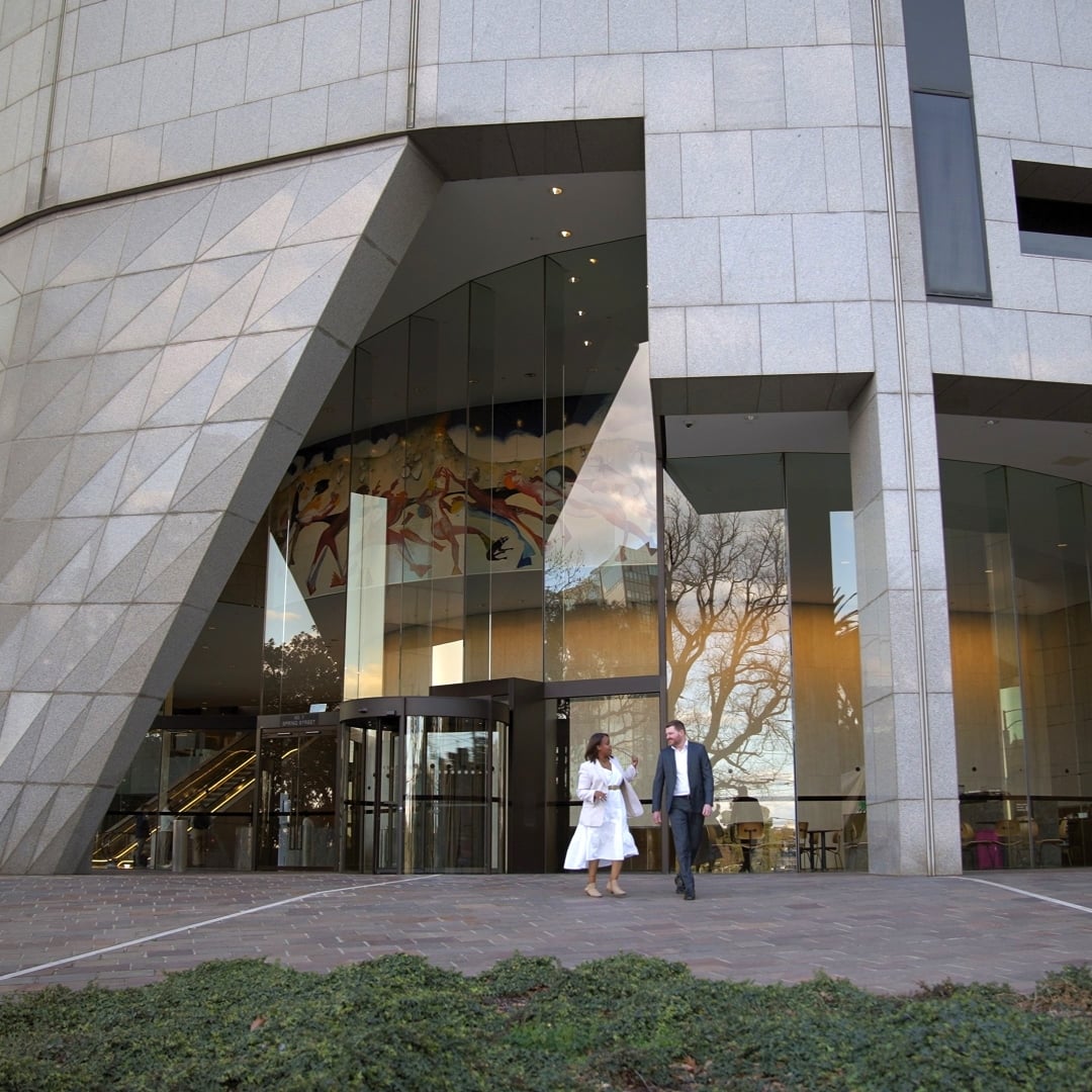 Two people walk out of a modern building with a geometric grey facade and large glass windows reflecting trees and a colourful mural.