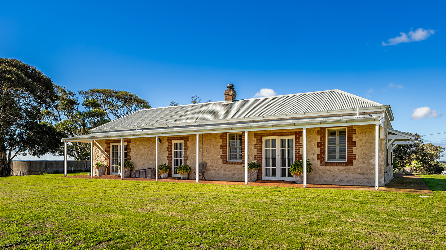 Historic stone farmhouse with a wraparound veranda and corrugated metal roof, set on a manicured lawn beneath a clear blue sky. 