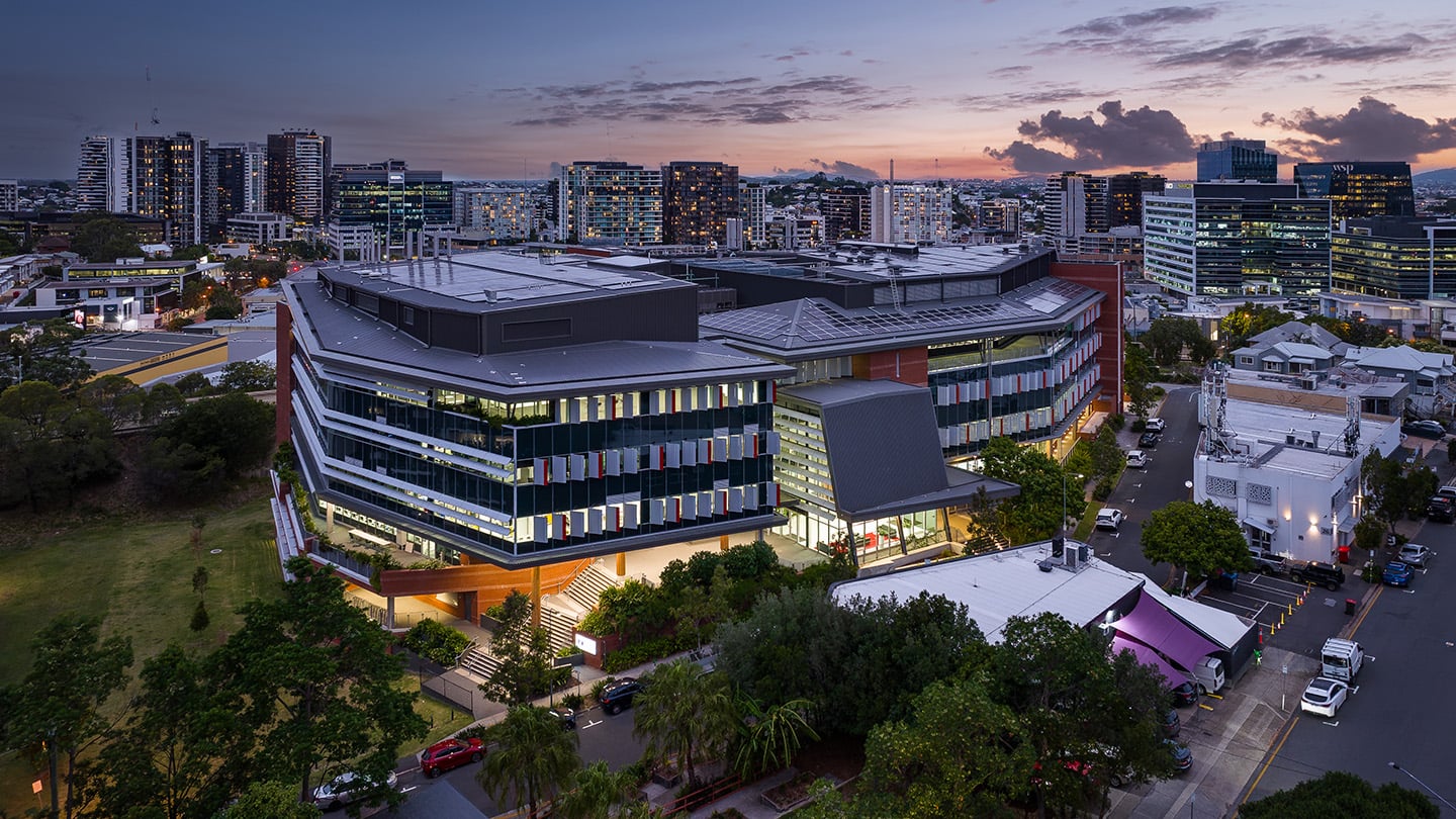 Aerial view of the Sullivan Nicolaides Pathology Central Laboratory, a modern multi-storey office and laboratory building illuminated at dusk, surrounded by trees, streets, and a dense urban skyline under a pink and blue evening sky. 