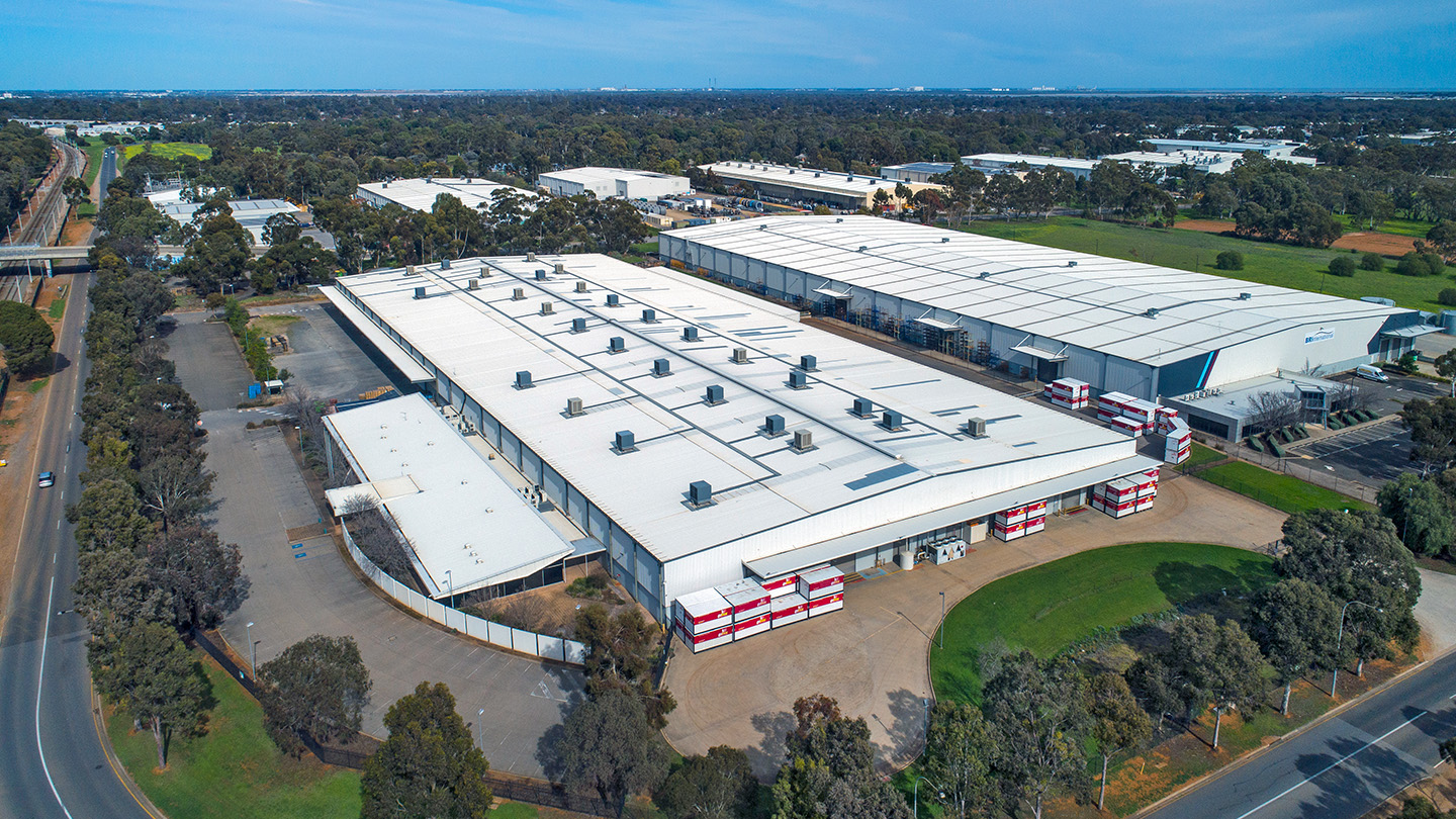 Aerial view of a large industrial warehouse complex with expansive, white-roofed buildings, loading and storage areas, surrounding roads and trees, situated within a broader industrial precinct in Adelaide. 