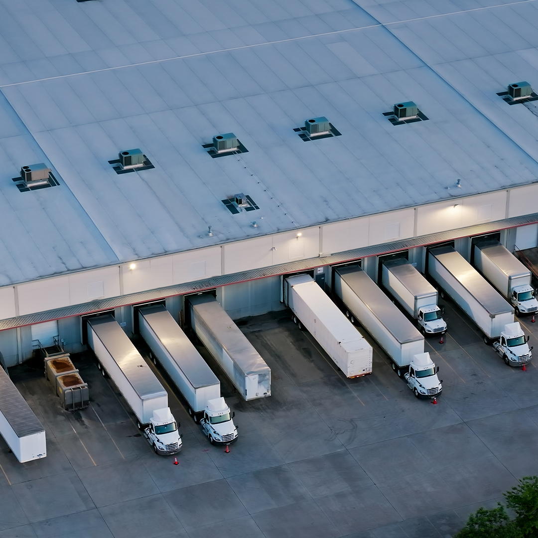 Aerial view of a warehouse loading dock with multiple semi-trailer trucks parked in bays along a large industrial building. 