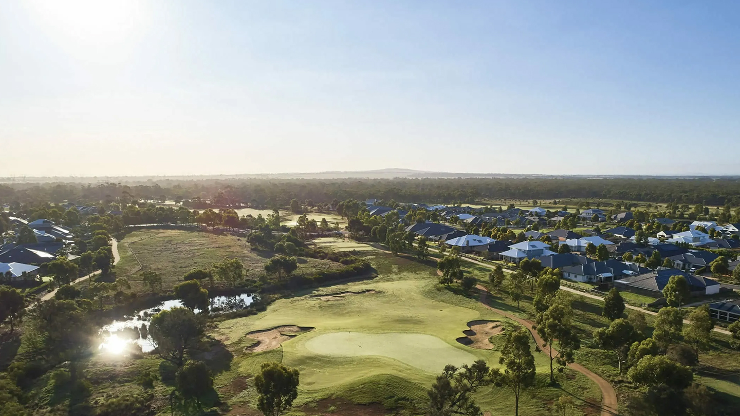 Expansive aerial view of green open space with rolling grassland, scattered trees and a water feature, bordered by a surrounding residential neighbourhood under a clear sky. 