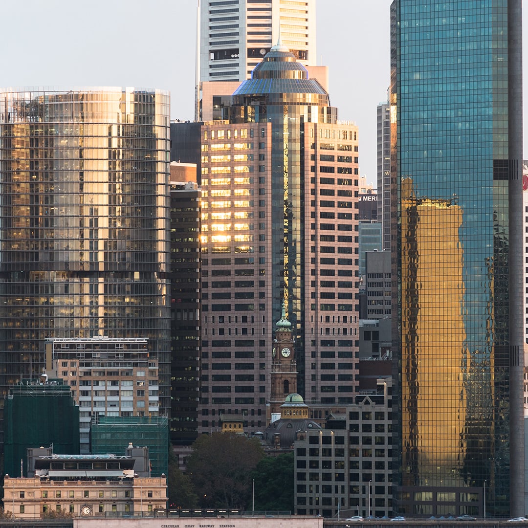 Sydney CBD skyline featuring modern office towers with reflective glass facades surrounding a historic clock tower at golden hour.