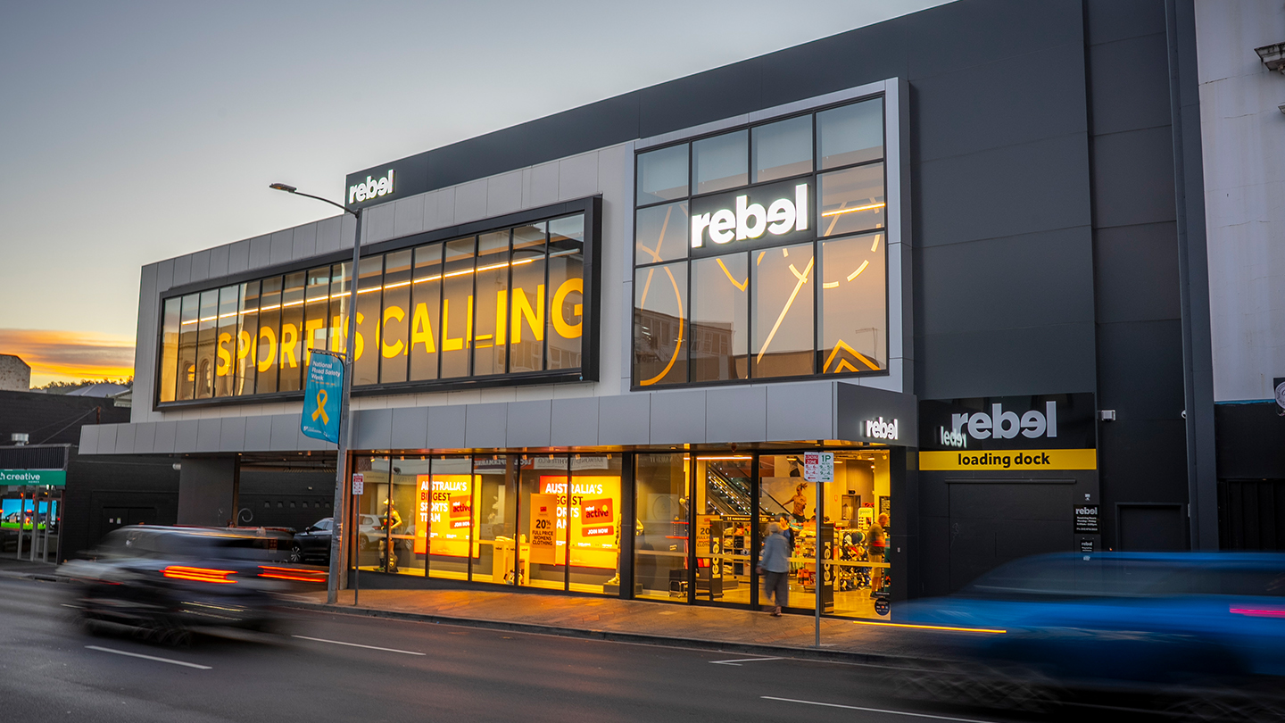 Street-level view of a modern Rebel sporting goods store at dusk, featuring illuminated storefront signage, large glass windows with bright interior displays, and blurred passing vehicles on the road in front. 