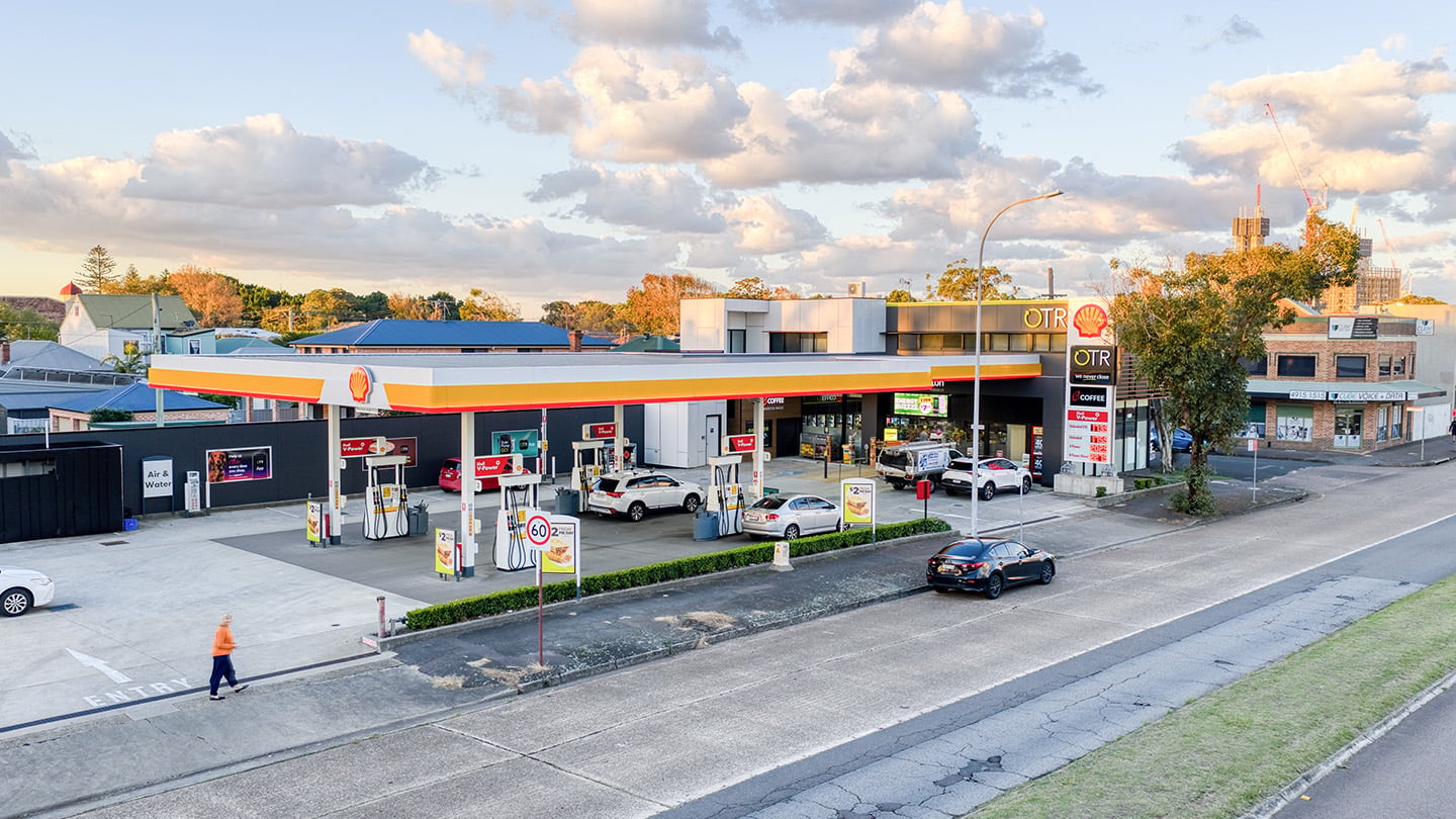 Aerial view of a Shell OTR service station and convenience store with multiple fuel pumps, parked vehicles, roadside signage, and surrounding suburban buildings under a partly cloudy sky. 