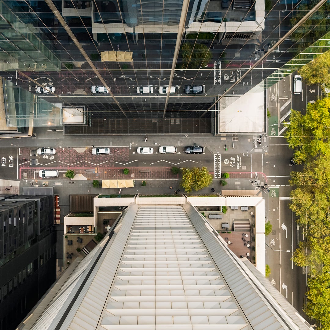 Top-down aerial view of a city street between modern high-rise buildings, with cars, bike lanes, sidewalks, and trees lining the road in a dense urban area.
