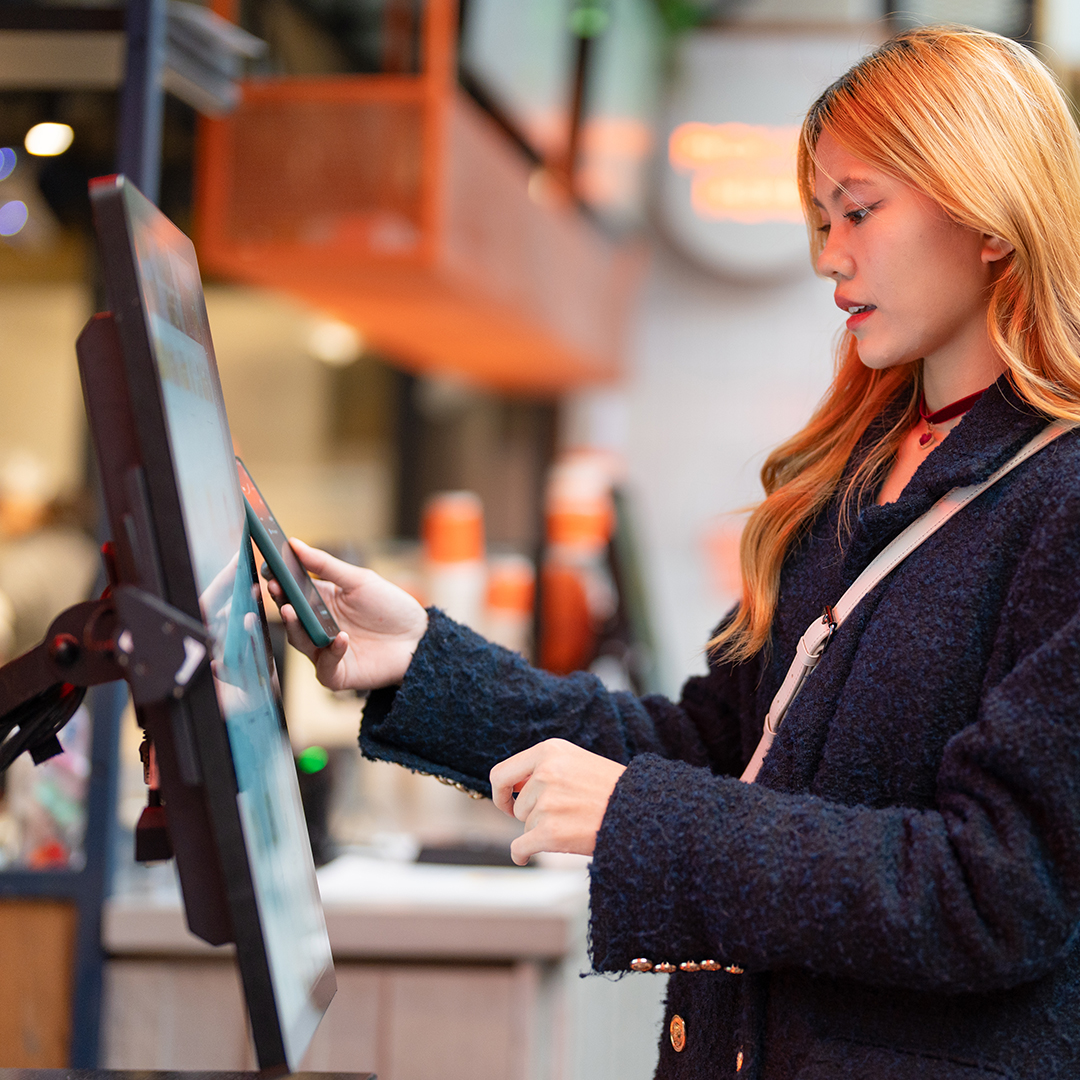 Person using a touchscreen self-service kiosk and smartphone in a modern quick-service environment. 
