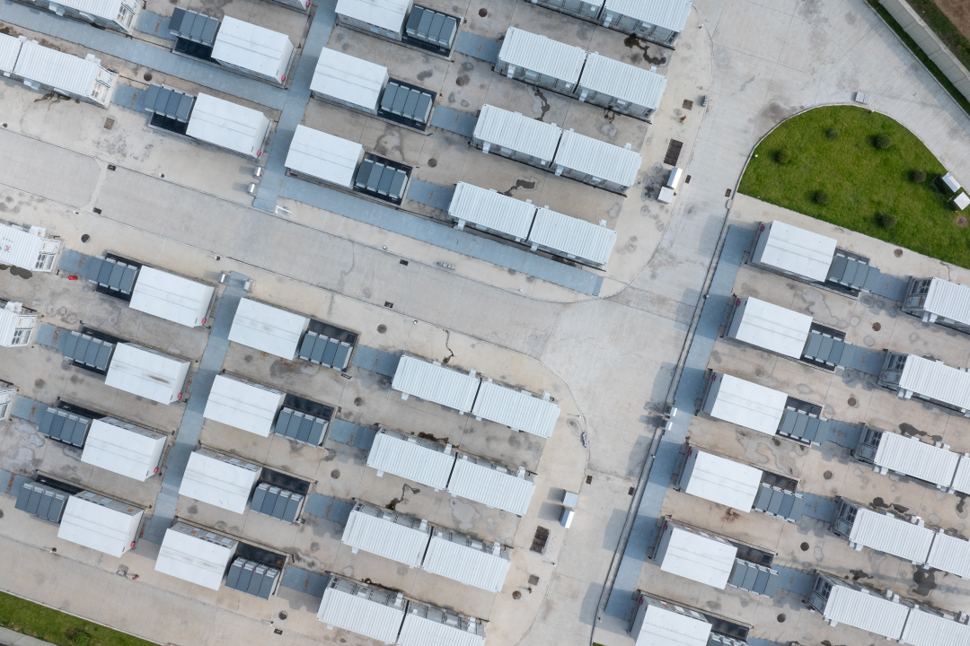 An aerial view captures numerous white modular buildings arranged in rows on a concrete surface, with a small patch of green grass visible in the top right.