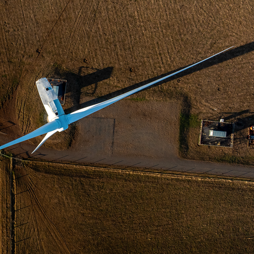An aerial shot captures a large wind turbine with two blades extended across farmland, casting long shadows.