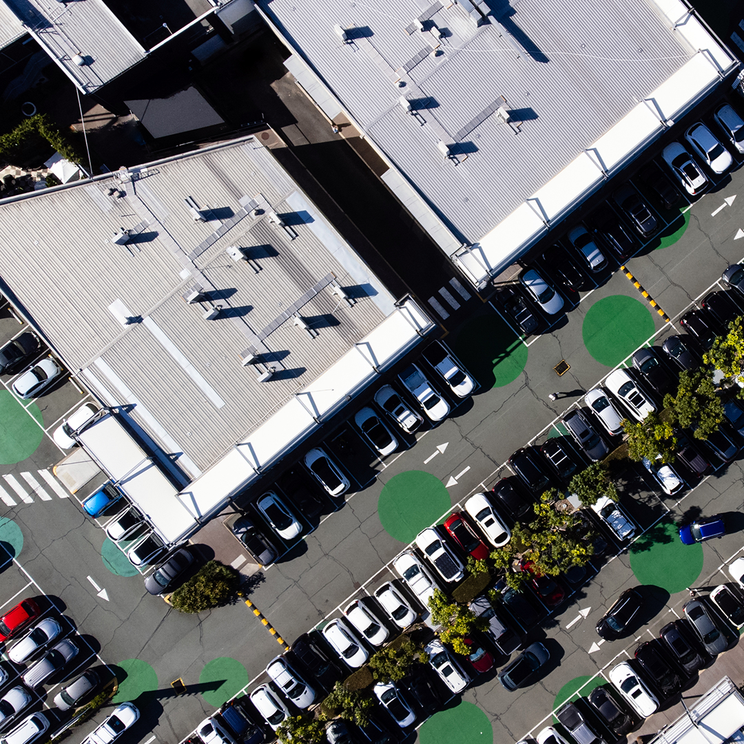An aerial view shows a sprawling retail complex with numerous cars parked in the sun-drenched lot, highlighted by green circles indicating parking spaces.