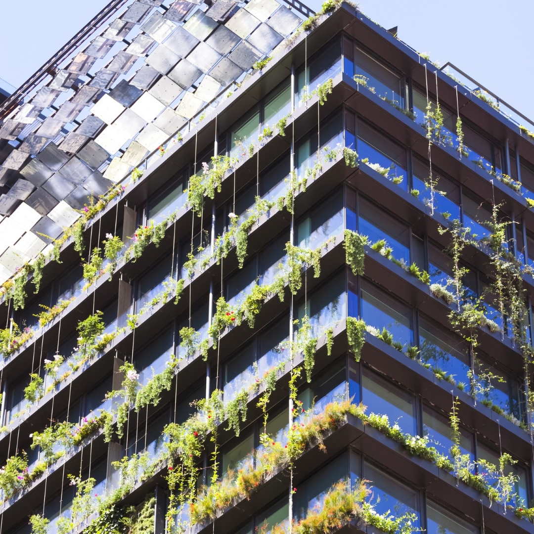 A modern building in a city is covered in lush green plants and vines, with a unique, textured roof reflecting the bright sky.