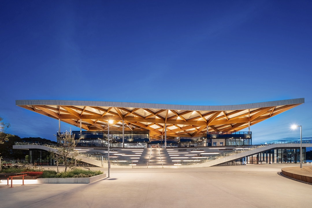 The new Sydney Fish Market building features a distinctive, curved, wooden lattice roof over a grand staircase leading to the entrance, illuminated at dusk under a clear blue sky.