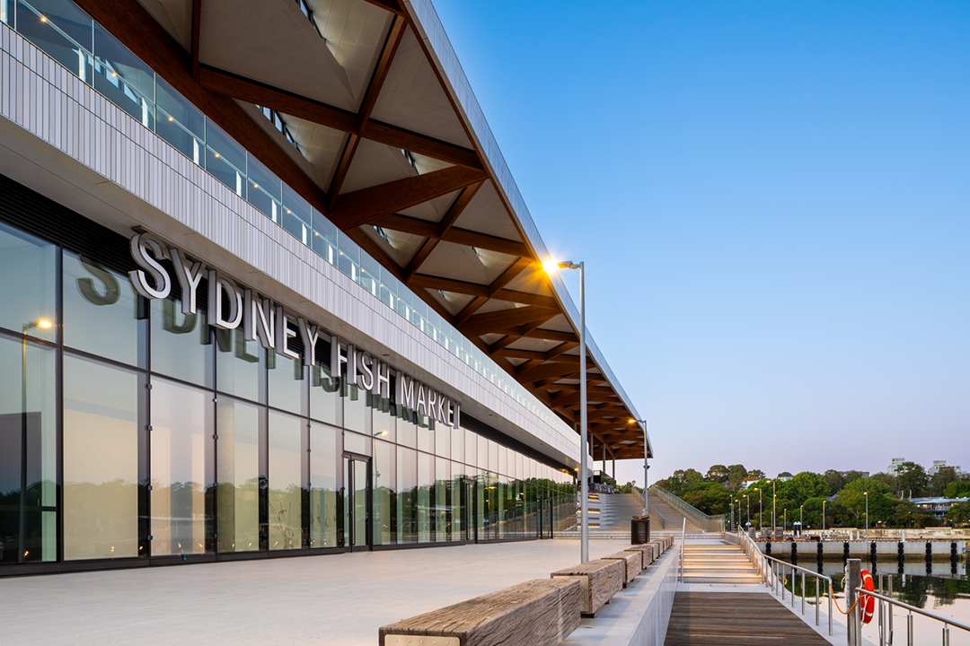 The new Sydney Fish Market building, featuring a large glass facade and a distinctive wooden-beamed roof, sits alongside a wide promenade with steps leading down to a waterfront pier.
