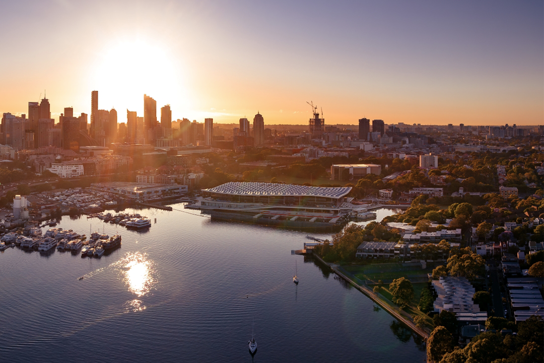 An aerial view of the Sydney skyline at sunset, with a large, modern building featuring a distinctive geometric roof situated on the water's edge in the foreground.