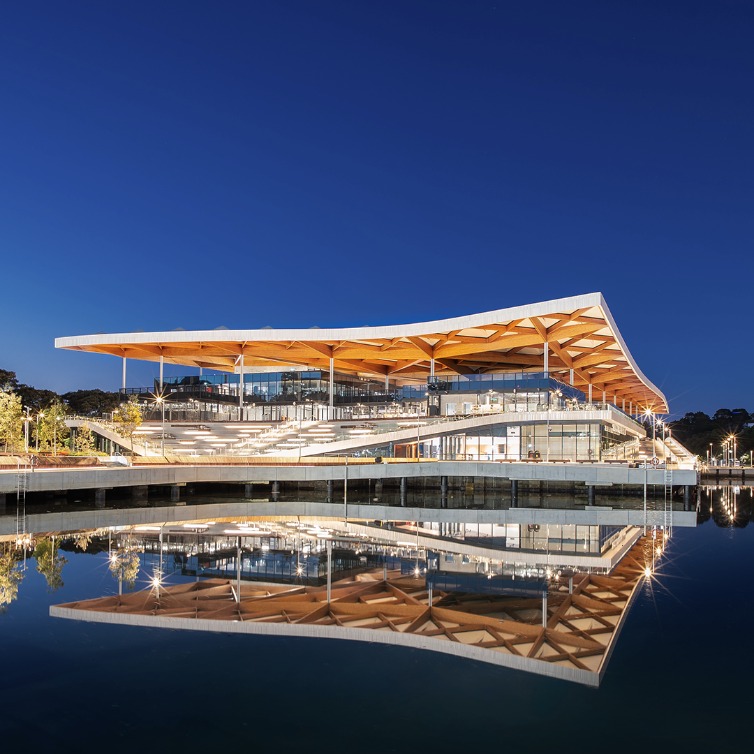 The striking new Sydney Fish Market building, featuring a distinctive curved roof and intricate wooden beams, is beautifully reflected in the water at twilight. 
