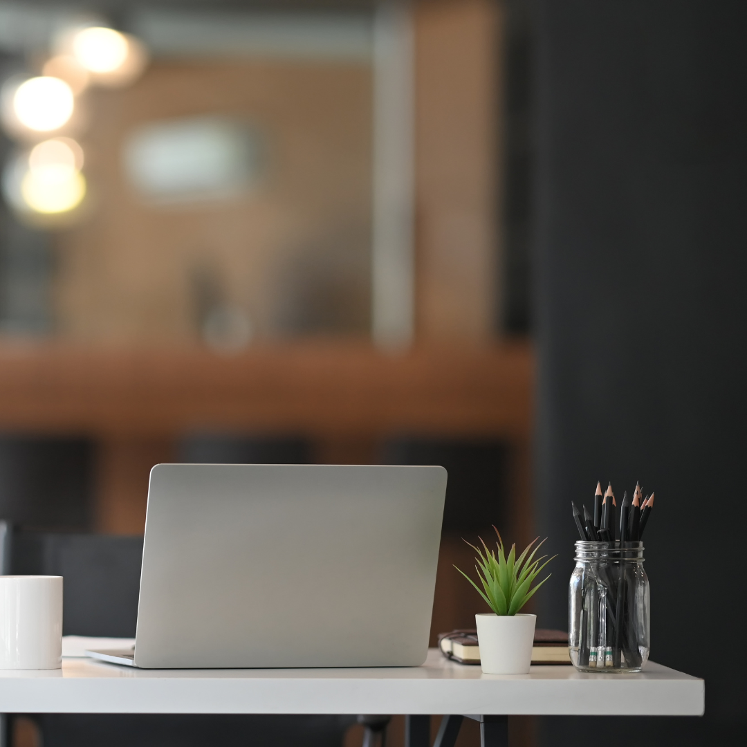 A white desk with a silver laptop, a white mug, a small potted plant, and a jar of pencils, set against a blurred background of an office space.