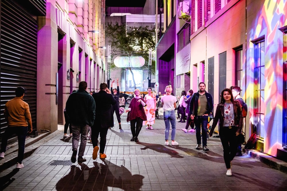 People gather in a vibrant urban alleyway illuminated by colorful lights, showcasing art installations and projections during the Flow and Glow event.