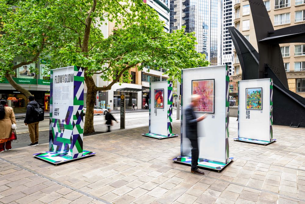 An outdoor art exhibition with vibrant geometric stands showcasing diverse artworks is set up on a city sidewalk, with blurred figures of people passing by.