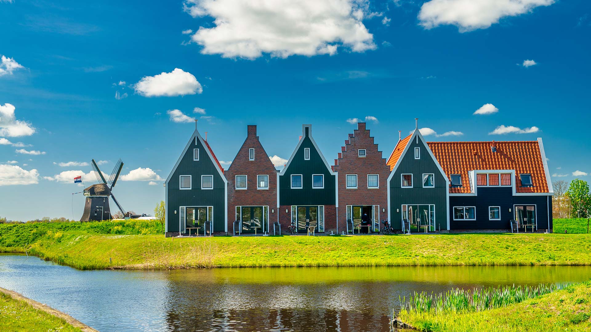 Vakantiepark met huisjes in Hollandse bouwstijl die aan het water staan en op de achtergrond een molen met een Nederlandse vlag..