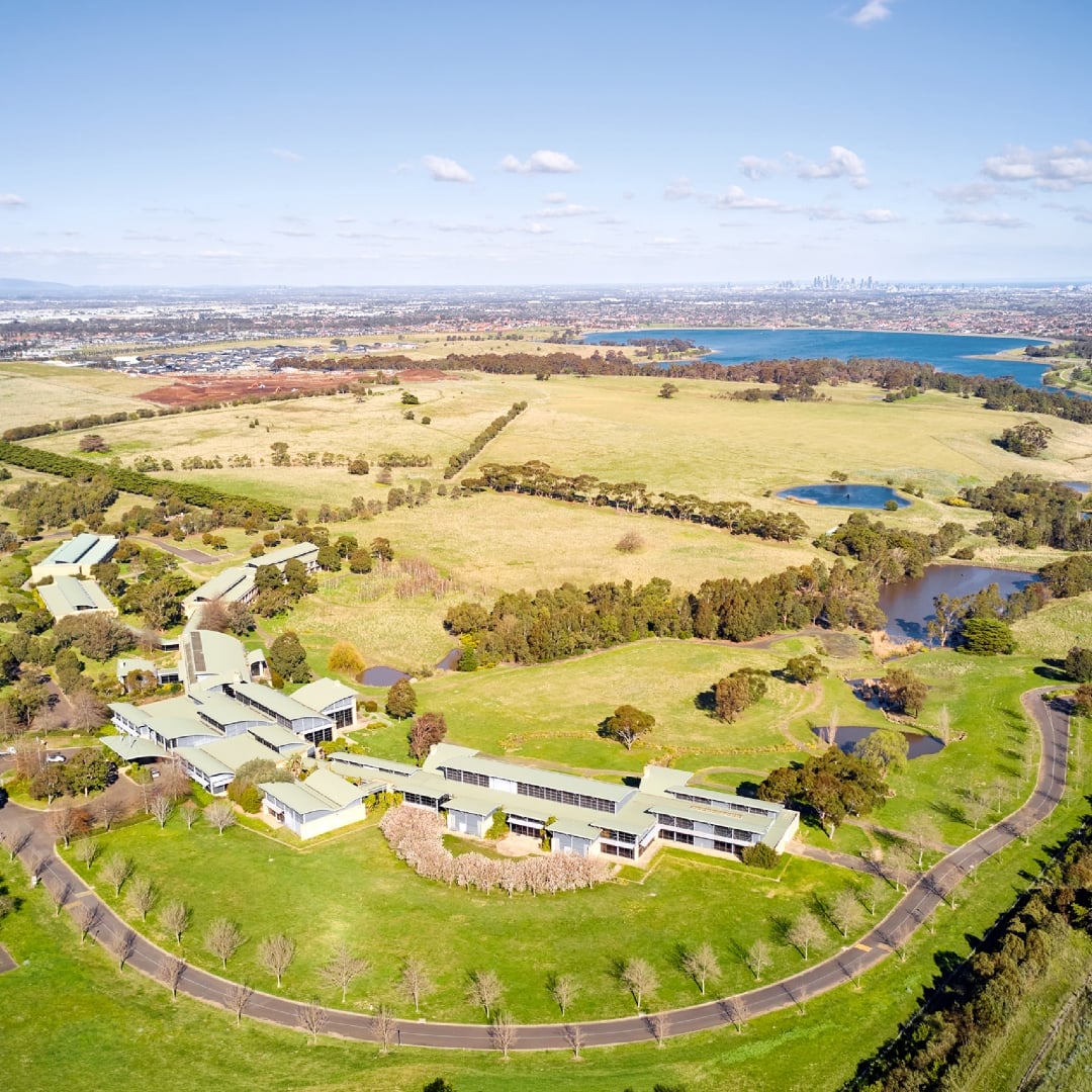 An aerial view captures a large, modern campus complex nestled within a sprawling landscape of rolling green paddocks and small dams, with a distant city skyline visible across a lake on the horizon.