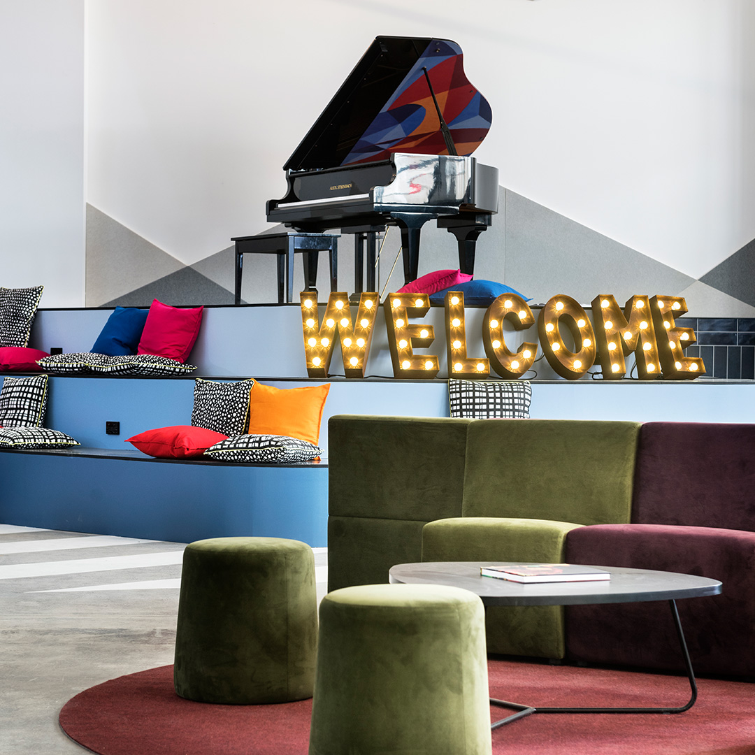 A colourful and inviting lounge area with a grand piano, plush seating, and a lit-up "WELCOME" sign.