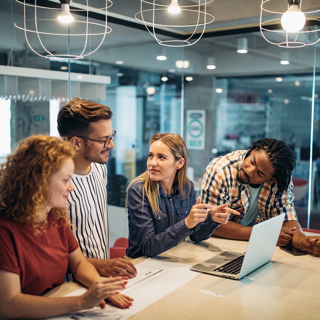 Four diverse young professionals collaborating around a laptop at a table in a modern creative office
