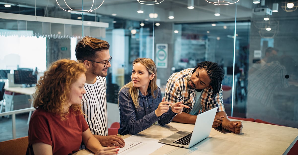 Four diverse young professionals collaborating around a laptop at a table in a modern creative office