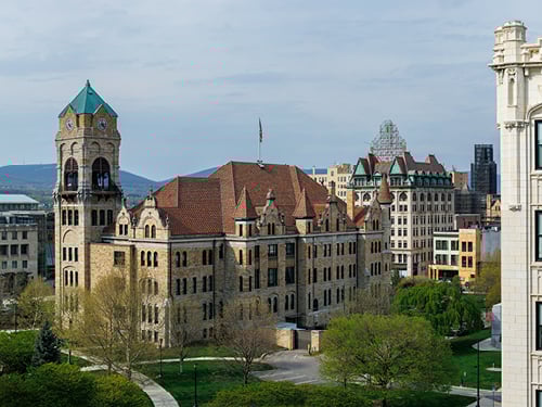 Historic stone building with a prominent green-roofed clock tower, surrounded by green parkland and other urban buildings under a clear sky.