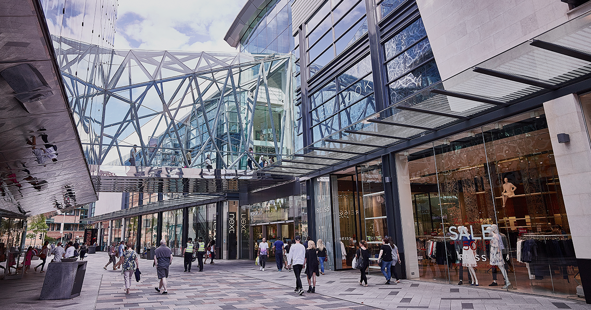 Photo of a modern shopping center's exterior, showcasing glass architecture and several retail stores. People are walking through the plaza.
