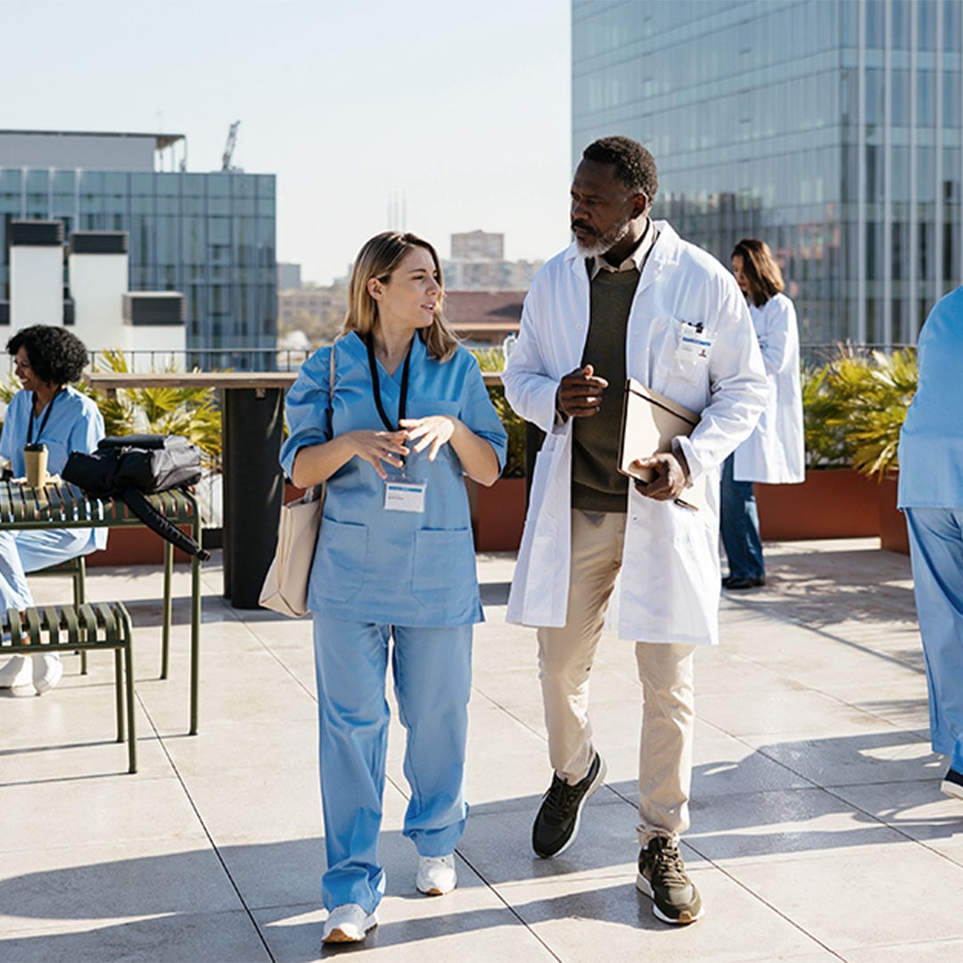 Doctors and nurses interacting on a rooftop with laptops and a wheelchair, surrounded by urban office buildings.
