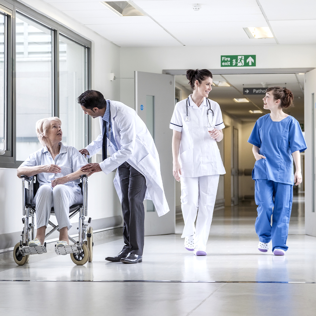 Modern hospital hallway with a doctor assisting a patient in a wheelchair and medical staff conversing, highlighting healthcare real estate.